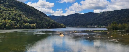 A serene river flows between lush green mountains under a partly cloudy sky. Reflections shimmer on the water's surface.