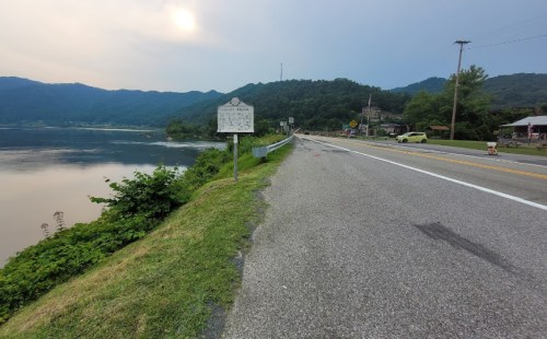 A quiet road alongside a river, with mountains in the background and a historical marker on the roadside.