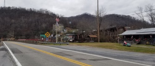 A rural road with a bridge, an American flag, and a small town in the background, surrounded by hills and overcast skies.
