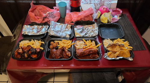 A table with several takeout containers filled with ribs, fries, and sides, alongside drinks and plastic bags.