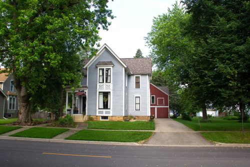 A charming two-story house with a blue exterior, red accents, and a well-kept lawn, surrounded by trees.
