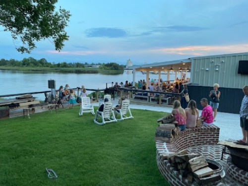 A riverside gathering with people enjoying a sunset, seated on lawn chairs and around a fire pit.