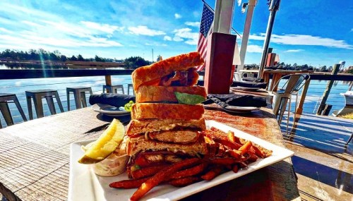 A towering sandwich with layers of ingredients, served with sweet potato fries and a pickle, against a scenic waterfront backdrop.
