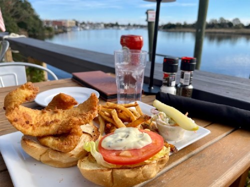 A plate of fried fish sandwich with fries, a pickle, and a view of a calm river in the background.
