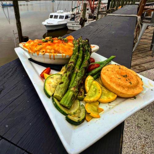 A plate of grilled vegetables, cornbread, and a bowl of macaroni and cheese, with a waterfront view in the background.