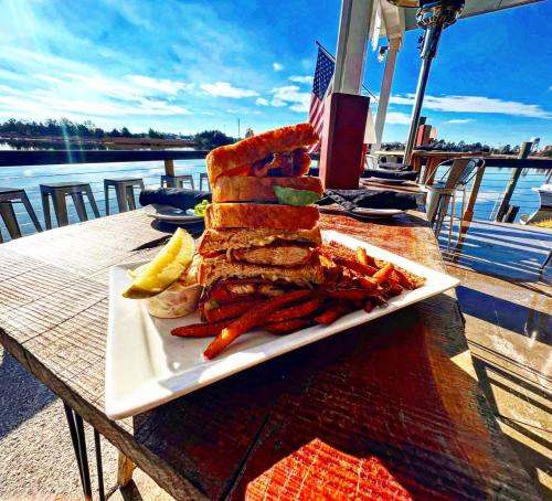 A towering sandwich with fries on a plate, set on a wooden table by a waterfront, under a clear blue sky.