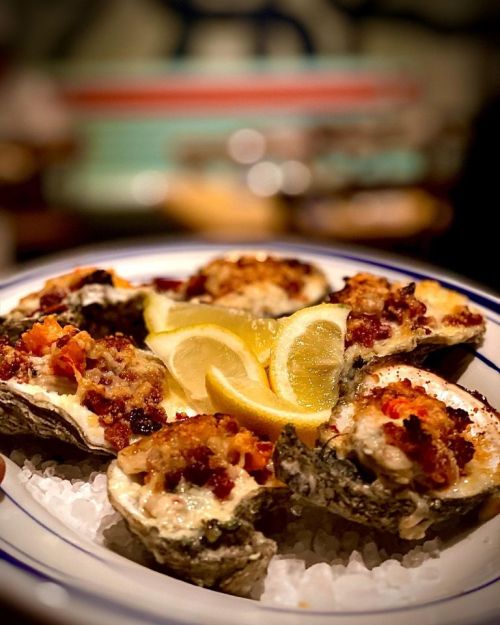 A plate of baked oysters topped with breadcrumbs and served with lemon wedges on a bed of ice.
