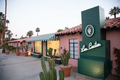 A colorful hotel exterior with cacti, a pool sign, and a neon "Los Cactus" sign, surrounded by palm trees.