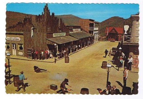 A vintage Western town scene with wooden buildings, people watching a shootout in the street, and barrels scattered around.