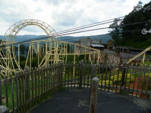 A roller coaster track winds through a scenic landscape with mountains in the background and a wooden fence in the foreground.