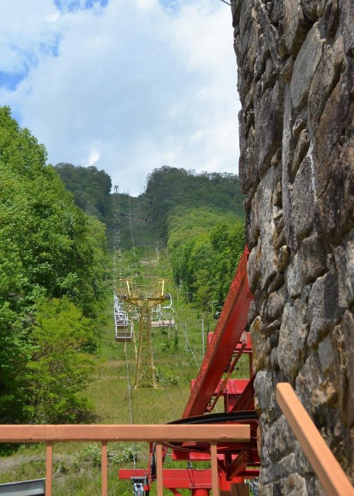 A ski lift ascends a green hillside, surrounded by trees and under a partly cloudy sky. Stone structure in the foreground.