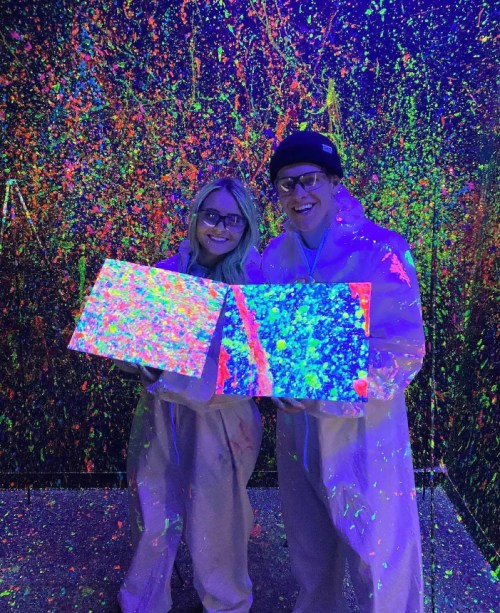 A couple in white suits holds glowing, colorful canvases in a vibrant, neon art space.