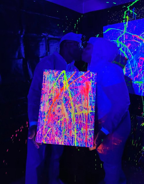 A couple in white outfits kisses while holding a brightly painted canvas under blacklight, surrounded by colorful splatters.