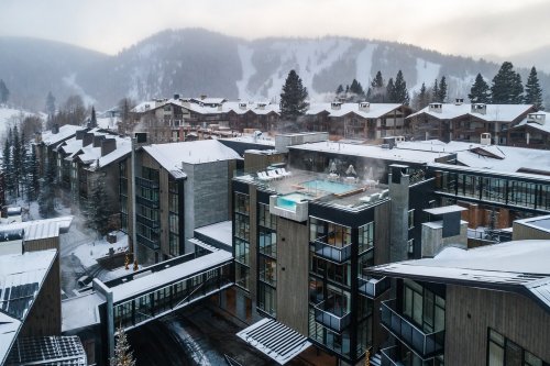 A snowy mountain resort with modern buildings and a rooftop pool, surrounded by trees and ski slopes in the background.