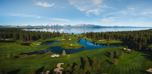 A panoramic view of a lush golf course surrounded by trees, lakes, and mountains under a clear blue sky.
