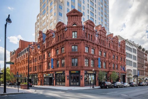 Historic red brick building at a city intersection, featuring shops on the ground floor and modern skyscrapers in the background.
