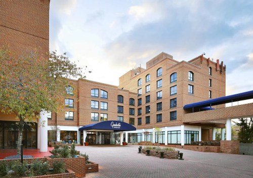 A modern brick hotel with a blue awning, surrounded by trees and a paved courtyard under a cloudy sky.