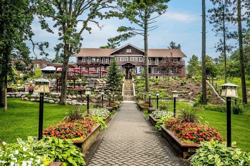 A scenic pathway lined with flowers leads to a large lodge surrounded by trees and landscaped gardens.