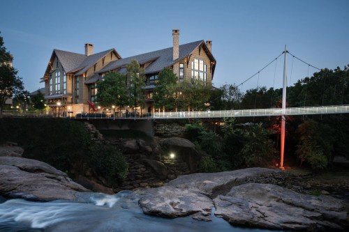 A scenic view of a rustic lodge by a river, featuring a pedestrian bridge and lush greenery at dusk.
