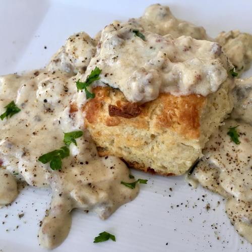 A fluffy biscuit topped with creamy sausage gravy, garnished with parsley and black pepper on a white plate.