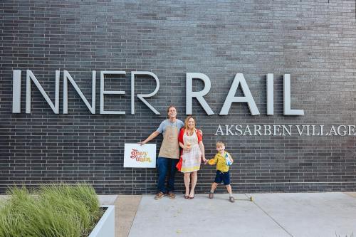 A family stands in front of a sign that reads "INNER RAIL" at Aksarben Village, holding a sign and autumn leaves.