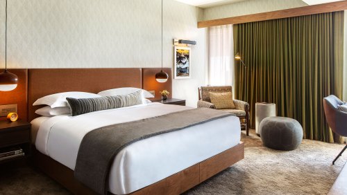 A modern hotel room featuring a king-sized bed, stylish decor, a cozy armchair, and large windows with green curtains.