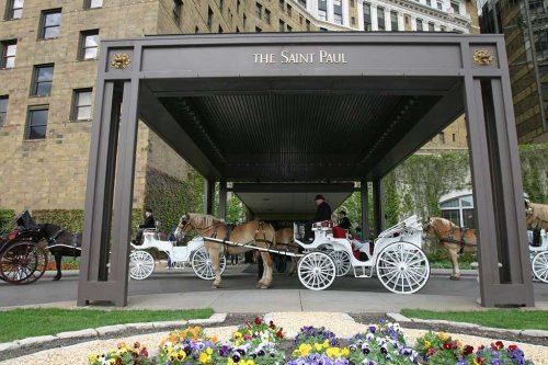 Carriage horses wait under a canopy at the Saint Paul hotel, surrounded by colorful flowers and historic architecture.