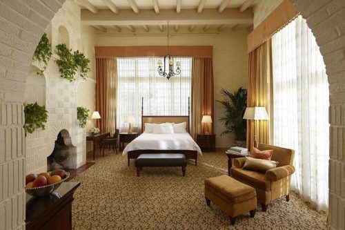 A spacious, elegantly decorated hotel room with a large bed, armchair, and bright windows, featuring warm tones and plants.