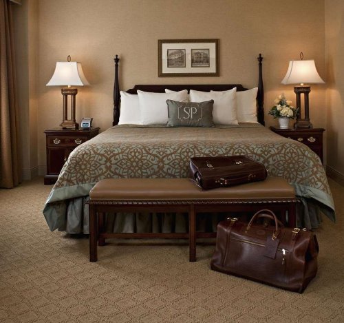 Cozy hotel room with a king bed, decorative pillows, a bench, and luggage on the floor. Warm, inviting decor.