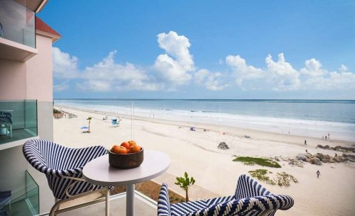 A balcony view overlooking a sandy beach with chairs and a bowl of oranges, under a bright blue sky.
