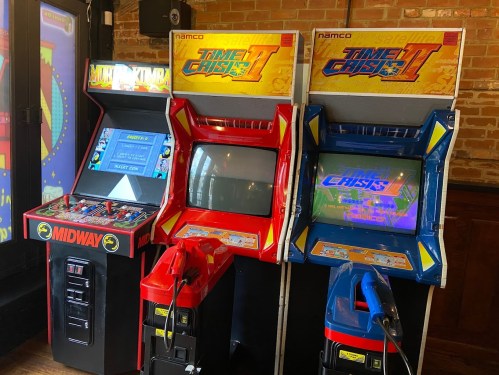 Three vintage arcade machines: "Mortal Kombat," "Time Crisis II" in red, and "Time Crisis II" in blue, set against a brick wall.