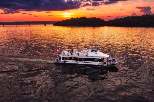 A boat cruises on a river at sunset, with vibrant orange and purple skies reflecting on the water.