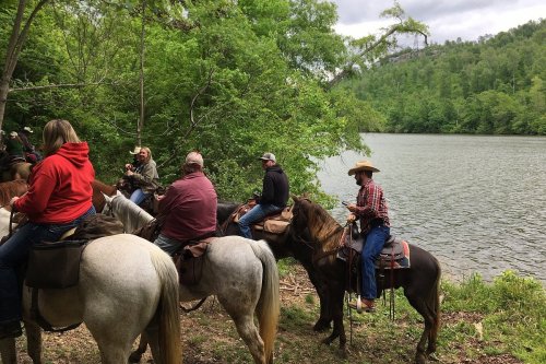 A group of people on horseback near a river, surrounded by lush greenery and hills in the background.