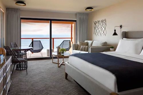 Cozy hotel room with a king bed, seating area, and balcony overlooking the ocean. Modern decor and natural light.
