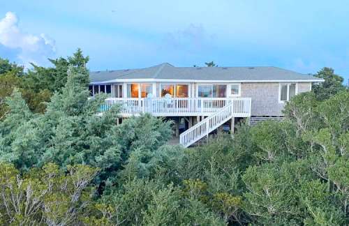 A coastal house with a deck, surrounded by trees, under a blue sky with soft clouds.