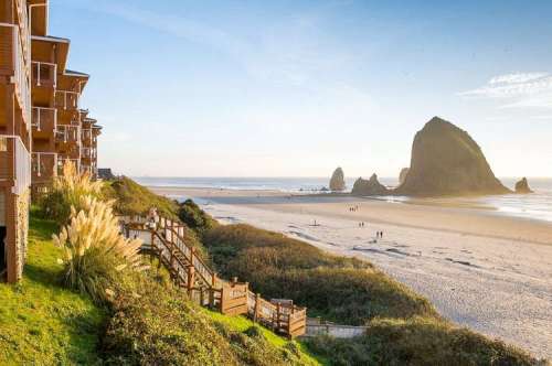 Coastal view with sandy beach, rocky formations, and wooden walkways, framed by buildings under a clear blue sky.
