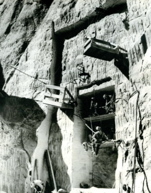 Black and white photo of workers on a rocky cliff, using scaffolding and equipment for excavation or construction.