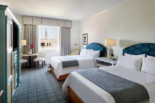 A bright hotel room with two beds, a small table, and a window showcasing a city view.