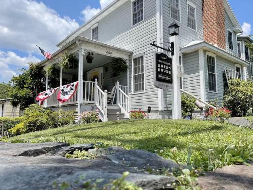 A charming guest house with a porch, American flags, and a sign, surrounded by lush greenery and a blue sky.