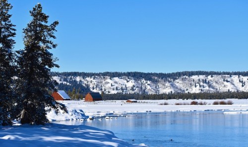 Snow-covered landscape with red barns, a river, and pine trees under a clear blue sky.