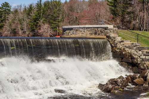 A waterfall cascades over a stone dam, surrounded by trees and a grassy area in a serene landscape.