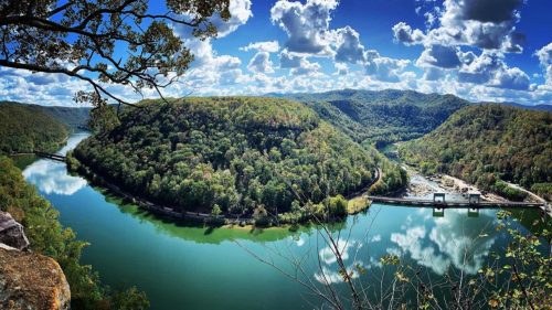 A panoramic view of a river winding through lush green hills under a bright blue sky with fluffy clouds.