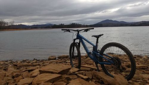 A blue mountain bike rests on rocky shore by a calm lake, with mountains and cloudy skies in the background.