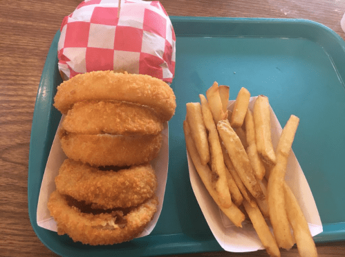 A teal tray holds a burger wrapped in red and white paper, alongside crispy onion rings and golden French fries.