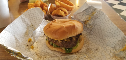 A cheeseburger with lettuce on a bun, served with onion rings and a drink in the background on a wooden table.