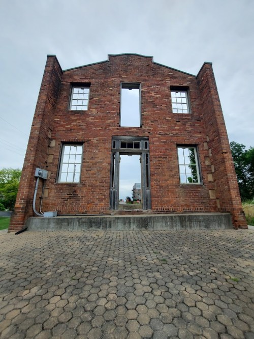 A brick building facade with empty windows and a doorway, set against a cloudy sky.