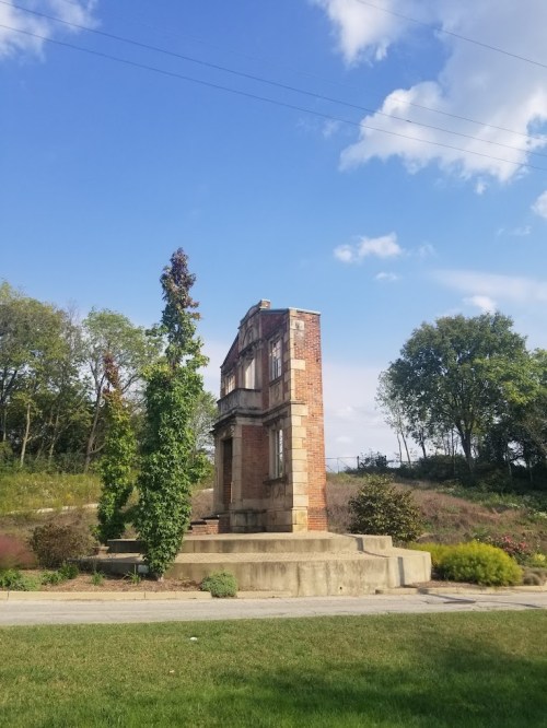 A partially standing brick structure surrounded by greenery under a blue sky with scattered clouds.