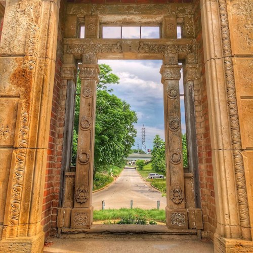 View through ornate stone doorway framing a tree-lined road under a cloudy sky.
