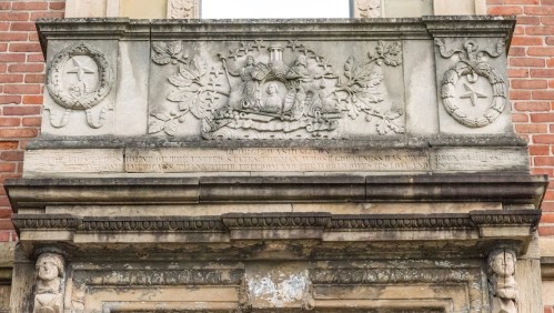 Stone relief featuring a coat of arms, decorative foliage, and inscriptions on a brick building facade.