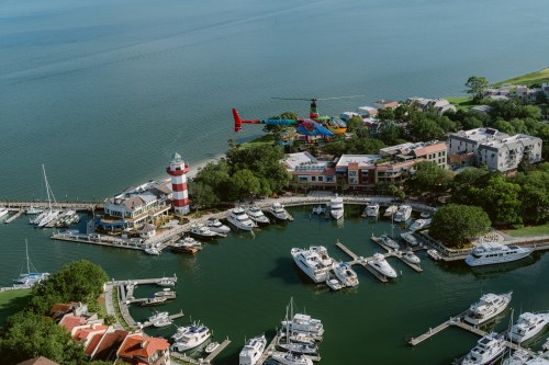 Aerial view of a marina with boats, a lighthouse, and a colorful helicopter flying above the water.